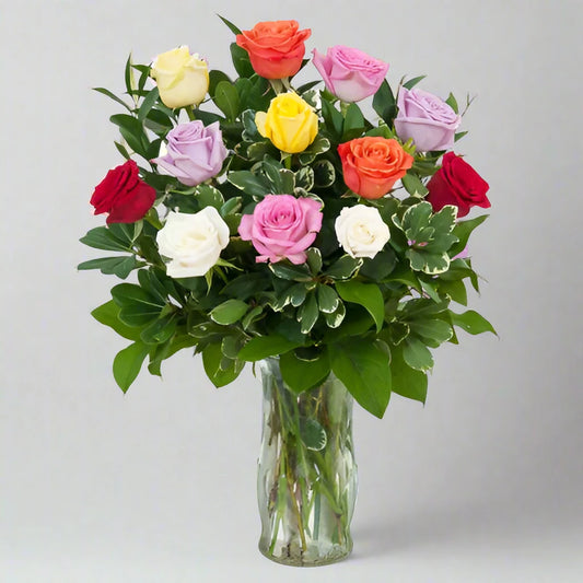 A beautiful bouquet of multicolored roses arranged in a clear glass vase. The roses come in various hues including red, yellow, orange, pink, white, and purple, interspersed with green leaves